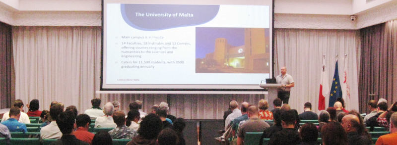 A lecture hall with people, a slide “The University of Malta” at the wall.